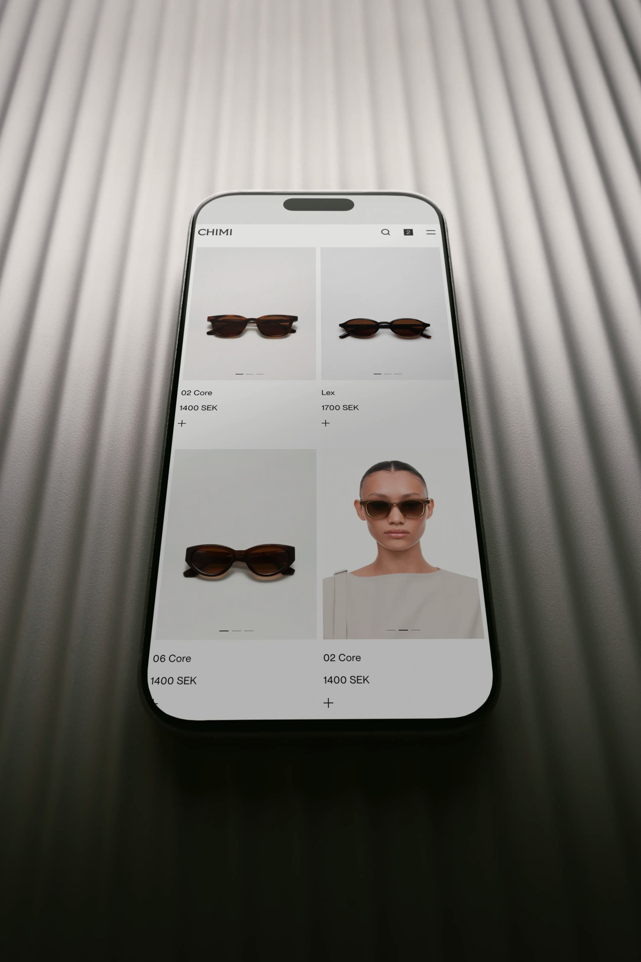 A smartphone displaying Chimis shopping experience, featuring various styles and prices. The phone rests on a ribbed, metallic surface.